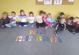 Dzieci siedzą obok napisu Dzień Przedszkolaka utworzonego samodzielnie z liter według odpowiedniego kodu.